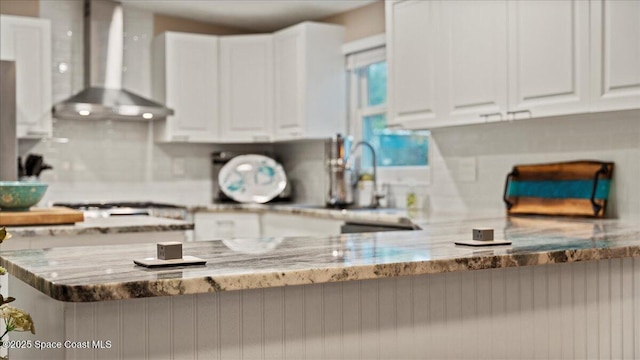 kitchen with light stone counters, wall chimney range hood, kitchen peninsula, and white cabinets