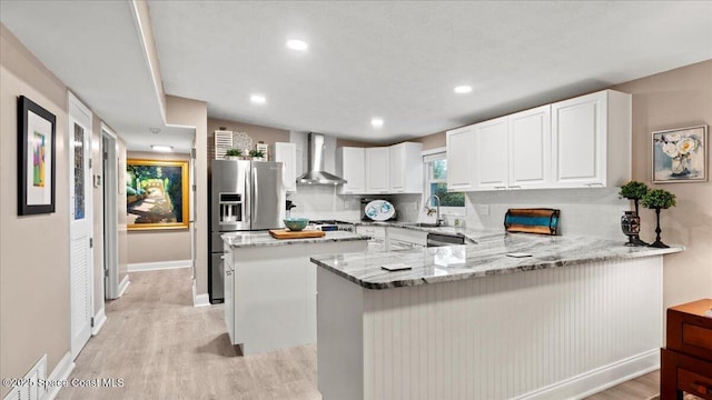 kitchen featuring stainless steel fridge, white cabinetry, light stone counters, kitchen peninsula, and wall chimney exhaust hood