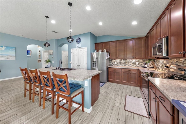 kitchen featuring vaulted ceiling, appliances with stainless steel finishes, decorative light fixtures, a breakfast bar area, and a center island with sink