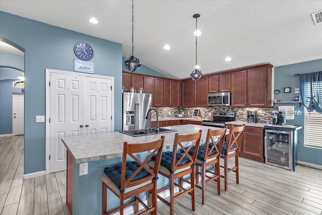 kitchen featuring lofted ceiling, stainless steel appliances, an island with sink, decorative light fixtures, and beverage cooler