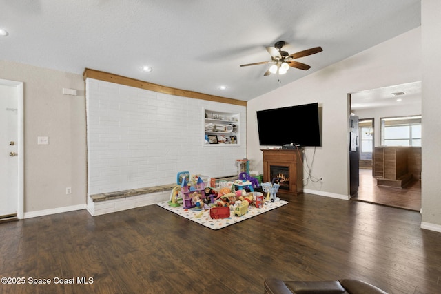 recreation room with ceiling fan, a warm lit fireplace, dark wood-type flooring, baseboards, and vaulted ceiling