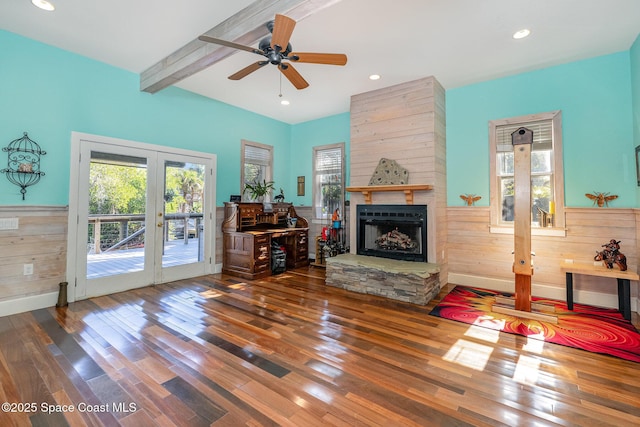 living room featuring a stone fireplace, wood-type flooring, wooden walls, ceiling fan, and beam ceiling