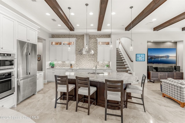 kitchen featuring wall chimney exhaust hood, decorative light fixtures, appliances with stainless steel finishes, an island with sink, and white cabinets