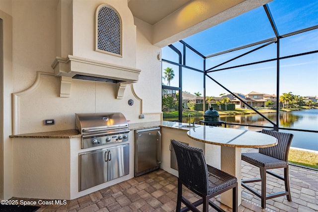 view of patio / terrace featuring a wet bar, a water view, glass enclosure, a grill, and an outdoor kitchen