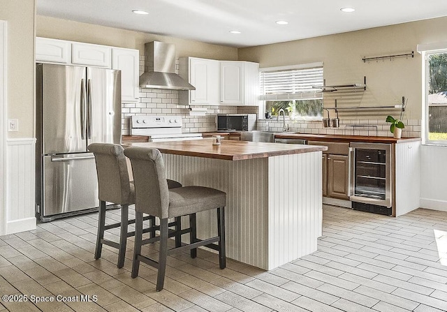 kitchen with wine cooler, wall chimney exhaust hood, white electric range, stainless steel refrigerator, and white cabinets