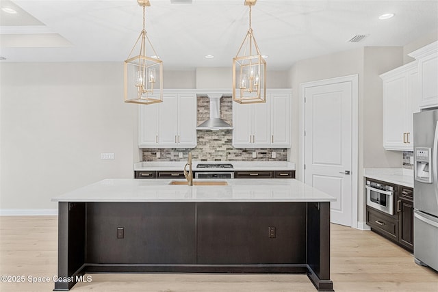 kitchen featuring white cabinets, decorative light fixtures, an island with sink, and wall chimney range hood