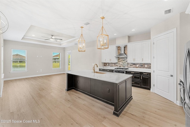 kitchen with wall chimney exhaust hood, sink, a center island with sink, pendant lighting, and white cabinets