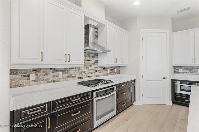 kitchen featuring white cabinets, light stone counters, stainless steel appliances, light wood-type flooring, and wall chimney exhaust hood