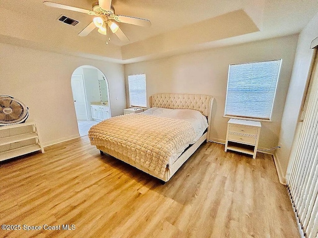 bedroom featuring ceiling fan, connected bathroom, a tray ceiling, and light hardwood / wood-style flooring