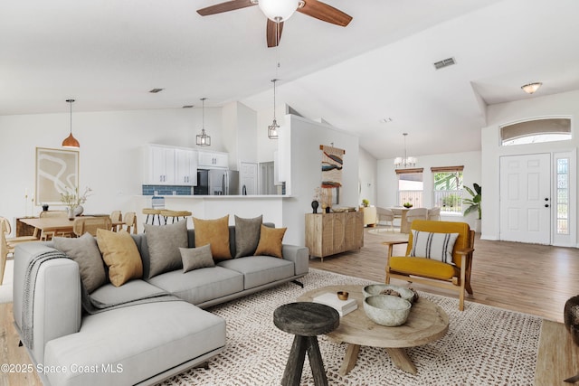 living area featuring ceiling fan with notable chandelier, light wood finished floors, visible vents, and high vaulted ceiling