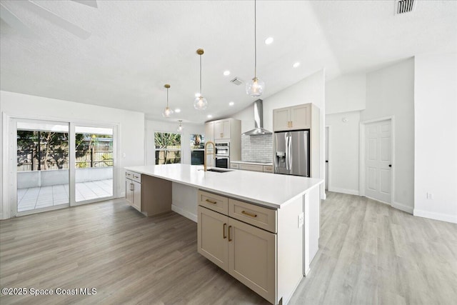 kitchen with light hardwood / wood-style flooring, hanging light fixtures, appliances with stainless steel finishes, a kitchen island with sink, and wall chimney range hood