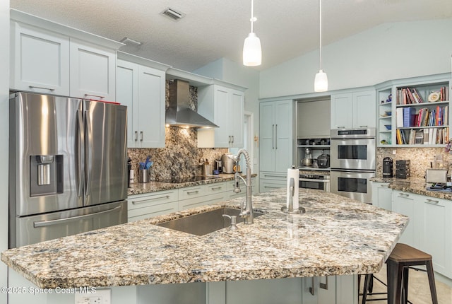 kitchen featuring wall chimney range hood, stainless steel appliances, an island with sink, and light stone countertops