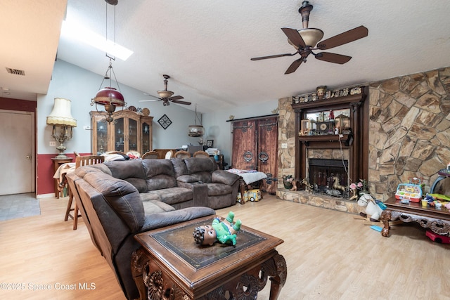 living room with a stone fireplace, vaulted ceiling, a textured ceiling, ceiling fan, and light hardwood / wood-style floors