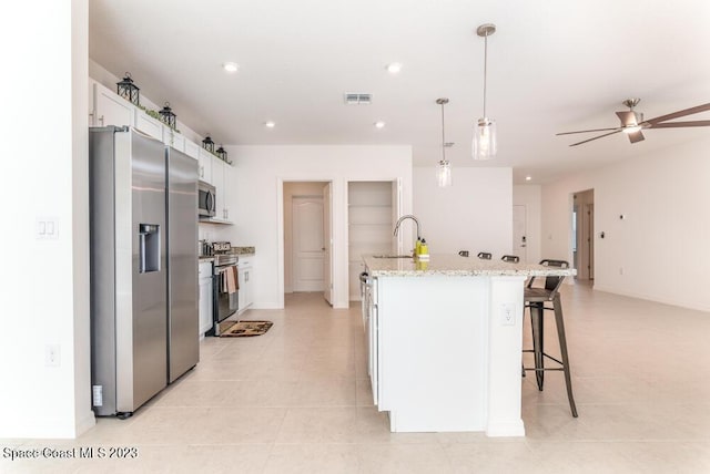 kitchen featuring light stone counters, hanging light fixtures, an island with sink, stainless steel appliances, and white cabinets