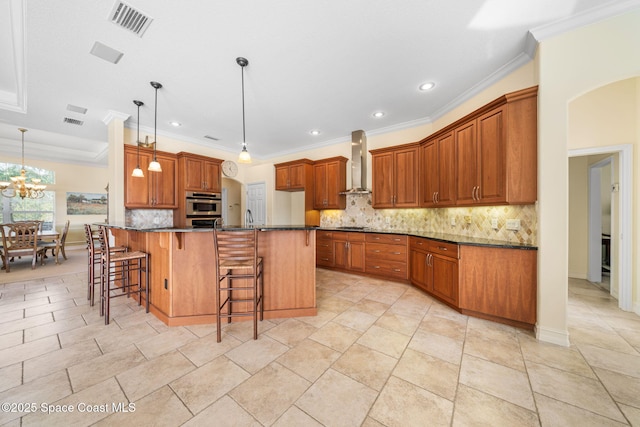 kitchen with a kitchen bar, hanging light fixtures, dark stone countertops, oven, and wall chimney range hood