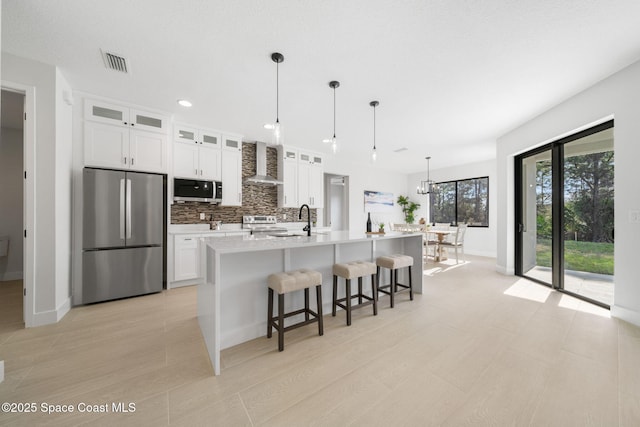 kitchen with pendant lighting, white cabinetry, wall chimney range hood, stainless steel appliances, and a center island with sink