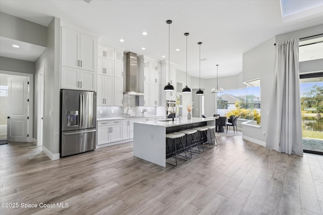 kitchen with white cabinetry, a kitchen breakfast bar, stainless steel refrigerator with ice dispenser, a center island with sink, and wall chimney exhaust hood