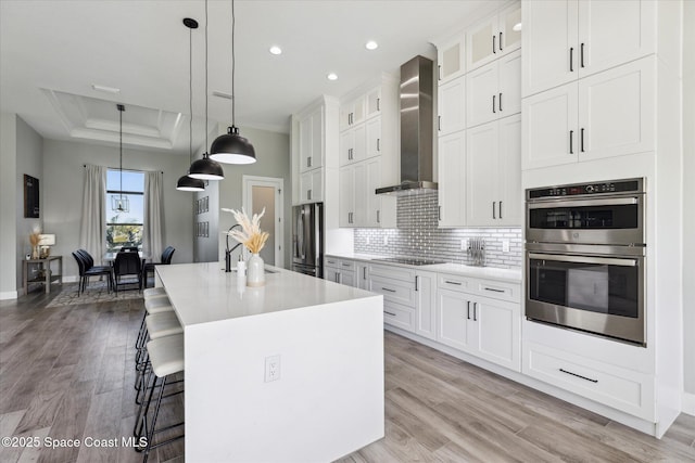 kitchen with wall chimney exhaust hood, white cabinetry, hanging light fixtures, a center island with sink, and appliances with stainless steel finishes