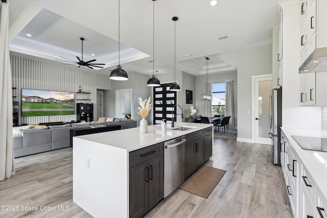 kitchen with sink, a raised ceiling, pendant lighting, stainless steel appliances, and a kitchen island with sink