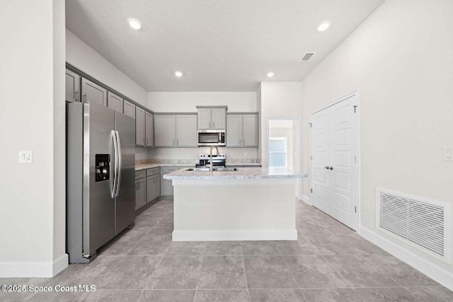 kitchen with sink, gray cabinetry, stainless steel appliances, light stone counters, and an island with sink