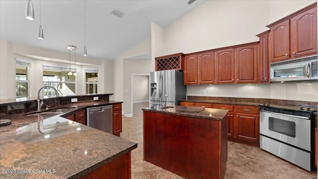 kitchen featuring a kitchen island with sink, stainless steel appliances, a sink, hanging light fixtures, and dark stone countertops