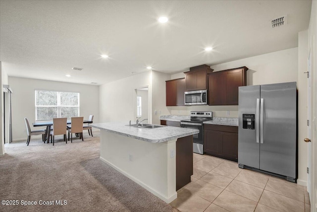 kitchen with sink, a kitchen island with sink, stainless steel appliances, dark brown cabinets, and light carpet