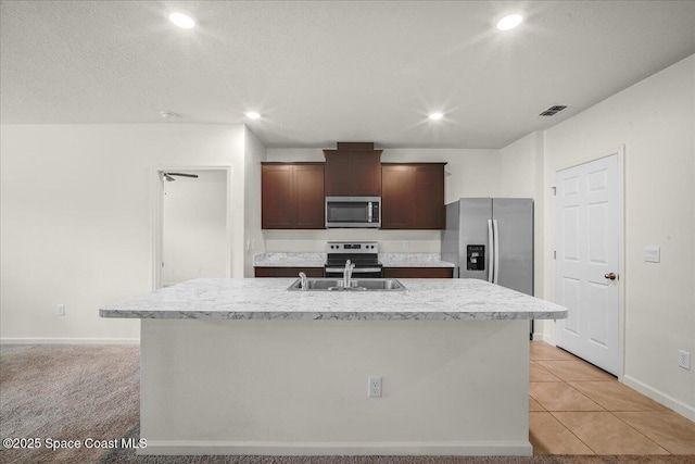 kitchen with dark brown cabinetry, sink, light tile patterned floors, stainless steel appliances, and a kitchen island with sink