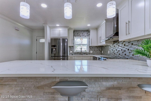 kitchen with wall chimney exhaust hood, hanging light fixtures, stainless steel fridge, light stone countertops, and white cabinets