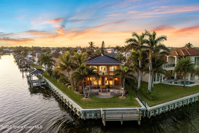 back house at dusk with a water view, a balcony, a patio area, and a lawn