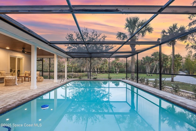 pool at dusk with ceiling fan, outdoor lounge area, glass enclosure, and a patio area