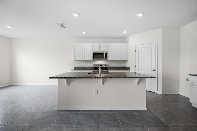 kitchen with white cabinets, stainless steel appliances, an island with sink, and dark stone countertops