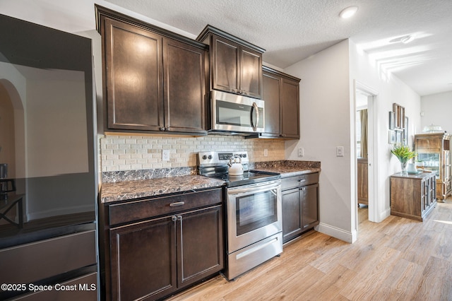 kitchen with tasteful backsplash, dark stone countertops, stainless steel appliances, dark brown cabinets, and light wood-type flooring