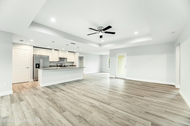 unfurnished living room featuring a raised ceiling, ceiling fan, and light wood-type flooring