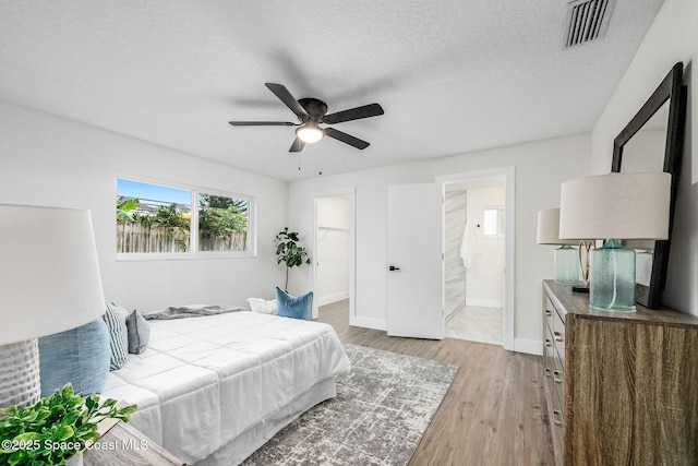 bedroom with ensuite bathroom, a spacious closet, a textured ceiling, light wood-type flooring, and ceiling fan