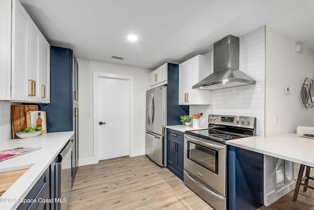 kitchen with appliances with stainless steel finishes, white cabinets, blue cabinetry, wall chimney range hood, and light hardwood / wood-style flooring