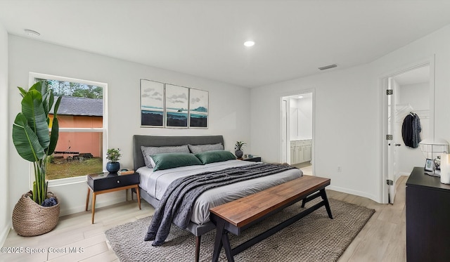 bedroom featuring light wood finished floors, baseboards, visible vents, ensuite bathroom, and a walk in closet