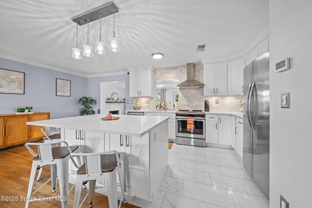 kitchen with a breakfast bar area, white cabinets, hanging light fixtures, stainless steel appliances, and wall chimney range hood