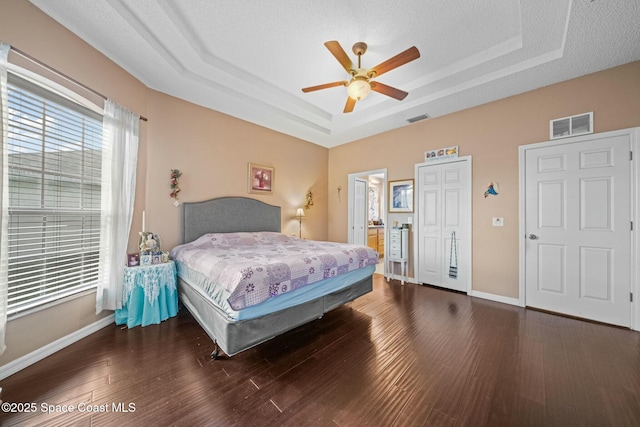 bedroom with dark wood-type flooring, ceiling fan, ensuite bathroom, and a tray ceiling