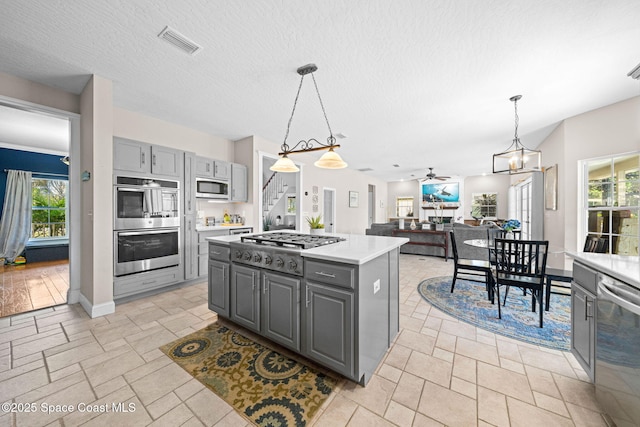 kitchen with gray cabinetry, hanging light fixtures, appliances with stainless steel finishes, a kitchen island, and ceiling fan with notable chandelier