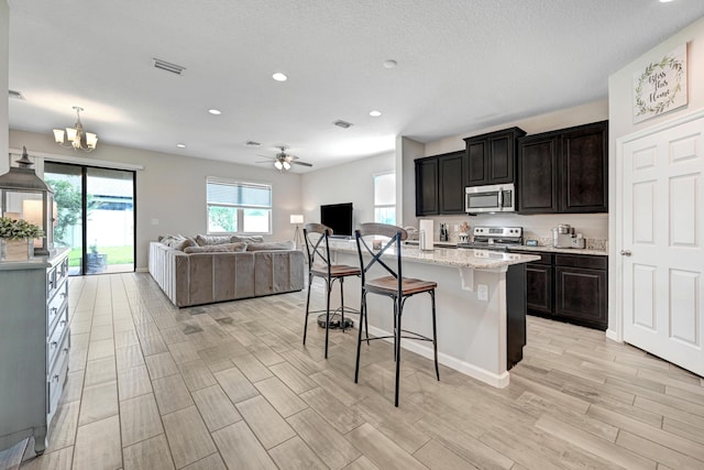 kitchen with a kitchen bar, light stone counters, appliances with stainless steel finishes, an island with sink, and ceiling fan with notable chandelier