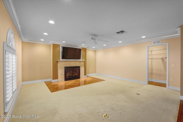 unfurnished living room with a tiled fireplace, crown molding, carpet floors, and ceiling fan