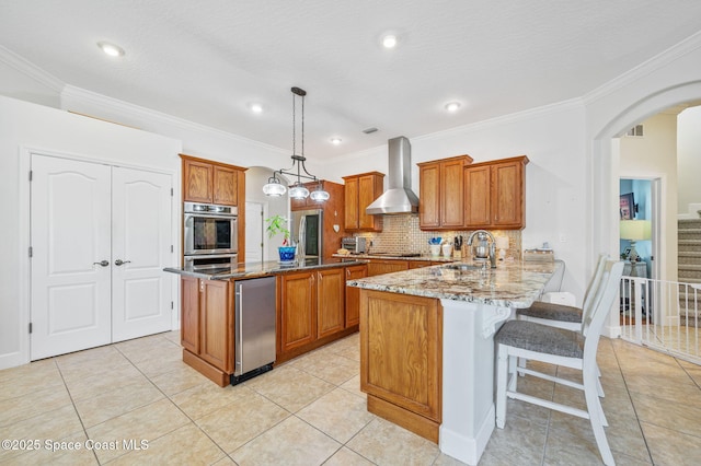 kitchen with a peninsula, a sink, hanging light fixtures, wall chimney range hood, and brown cabinets