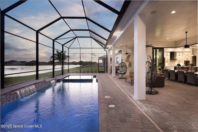 pool at dusk with pool water feature, a water view, a lanai, ceiling fan, and a patio