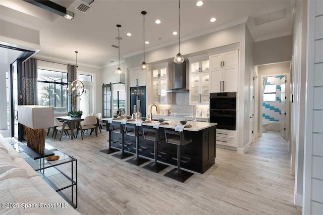 kitchen featuring decorative light fixtures, white cabinetry, an island with sink, black appliances, and wall chimney range hood