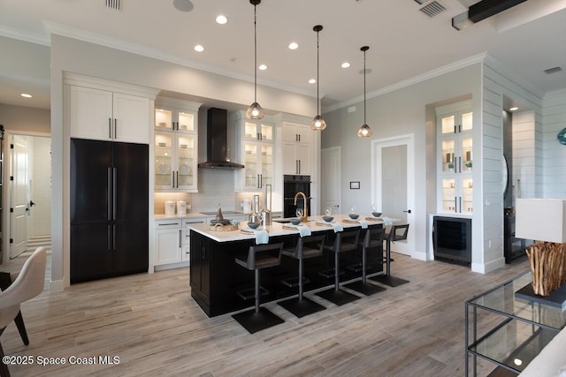 kitchen with decorative light fixtures, white cabinetry, a kitchen island with sink, black appliances, and wall chimney exhaust hood