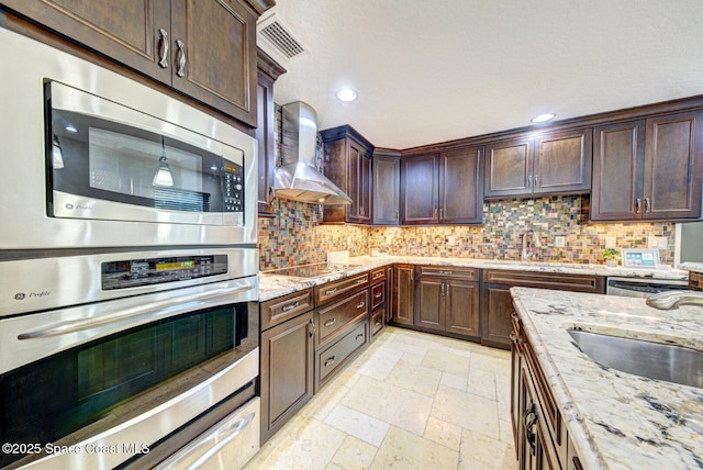 kitchen featuring dark brown cabinetry, light stone counters, wall chimney range hood, stainless steel appliances, and backsplash