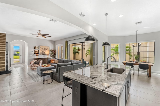 kitchen featuring sink, hanging light fixtures, light tile patterned floors, light stone countertops, and a center island with sink