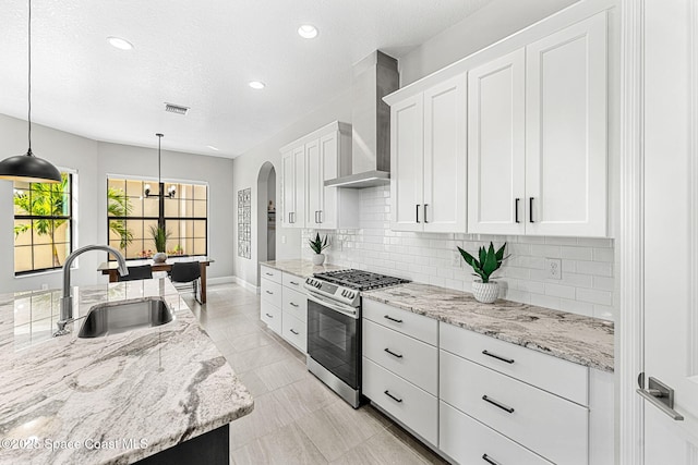 kitchen featuring wall chimney exhaust hood, sink, gas stove, white cabinetry, and pendant lighting