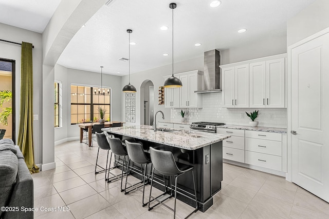 kitchen with decorative light fixtures, sink, white cabinets, wall chimney range hood, and a center island with sink