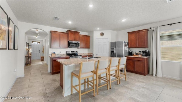 kitchen featuring a breakfast bar, a center island with sink, a textured ceiling, appliances with stainless steel finishes, and light stone countertops